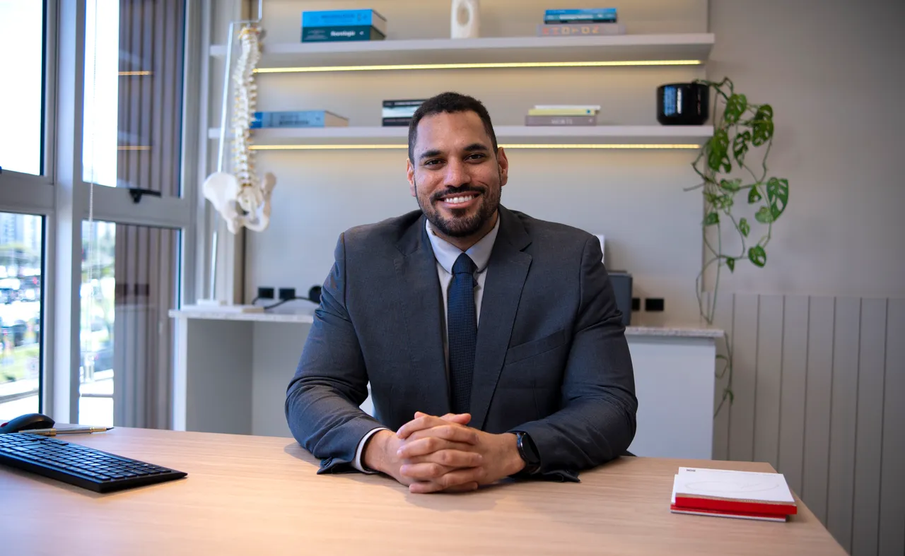 Dr. Elson Shockness sorrindo, sentado à mesa do consultório, de terno e gravata